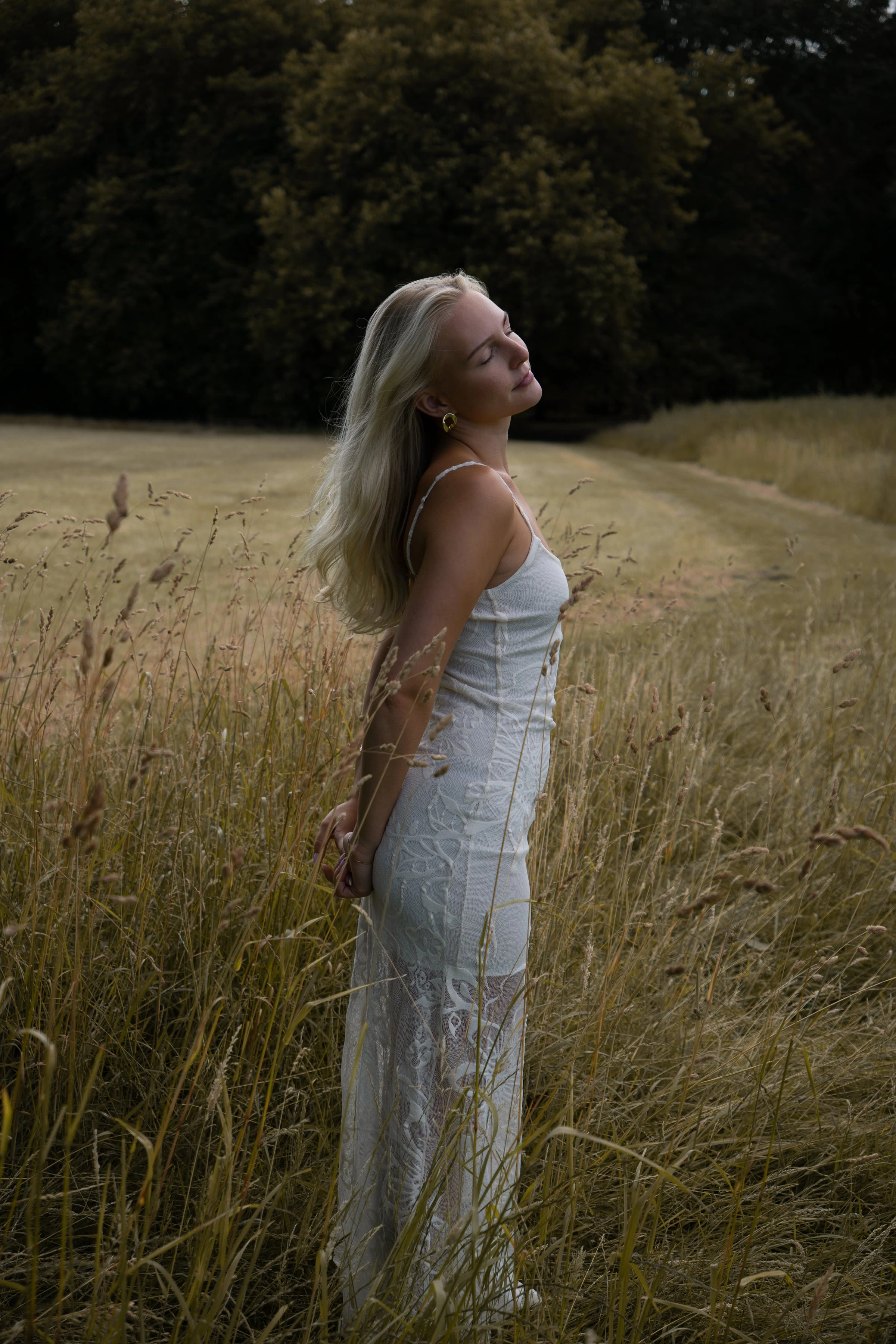 Vrouw in witte kanten jurk staat met gesloten ogen in een veld met lang gras