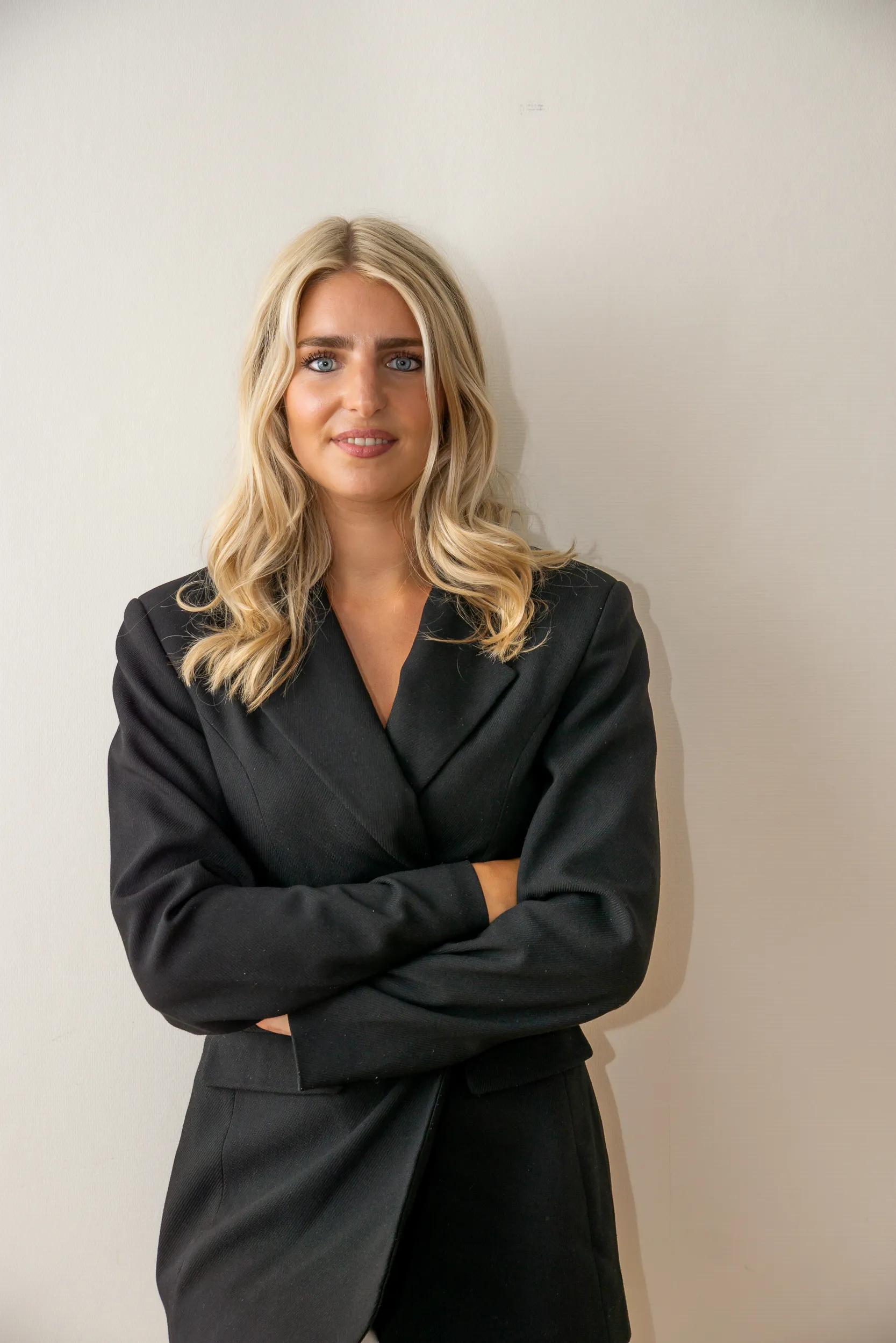 Vrouw met blonde golvende haren in zwarte blazer, armen over elkaar, warme glimlach tegen een lichte muur